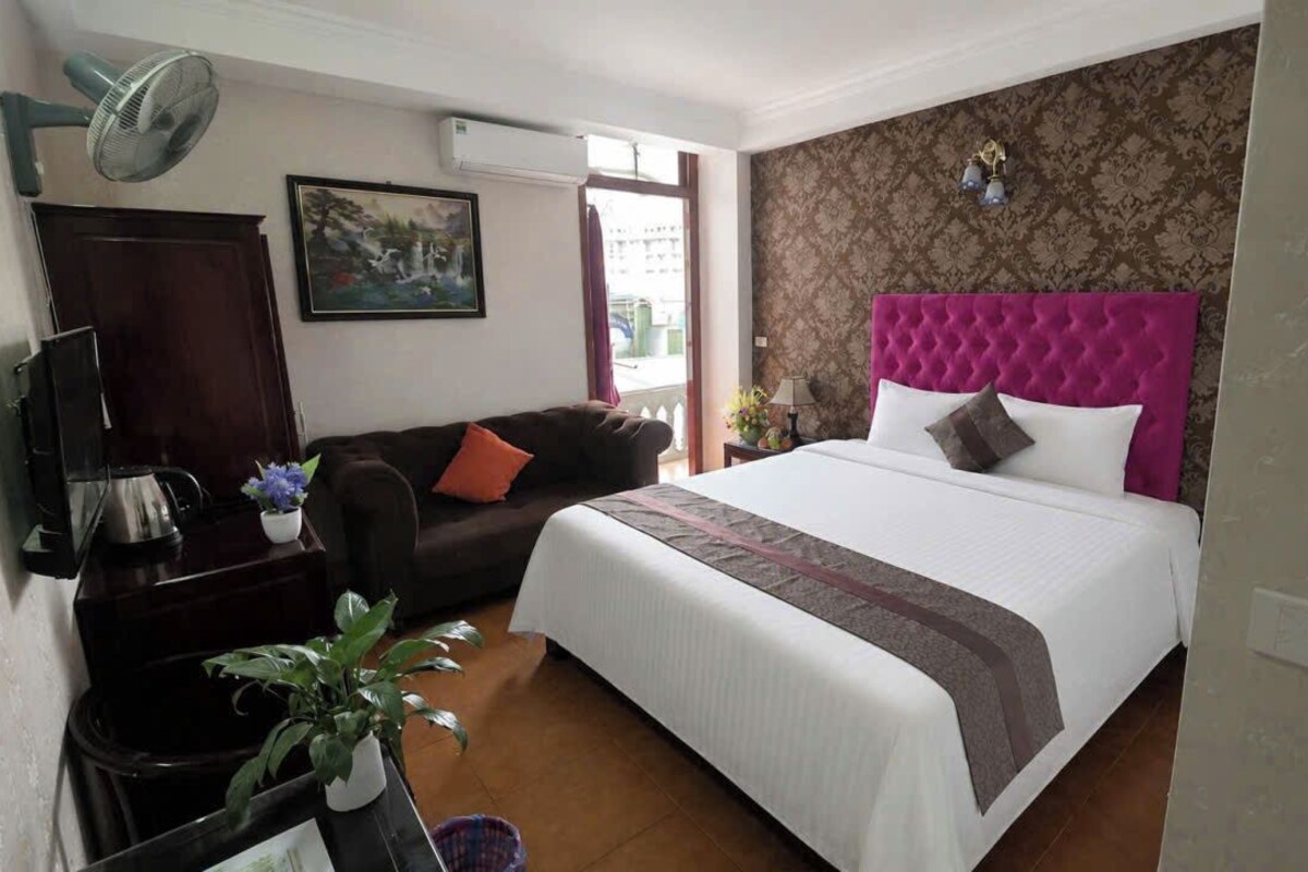 A hotel room with a bed, a sofa, a desk with a lamp, a plant, and a painting on the wall.