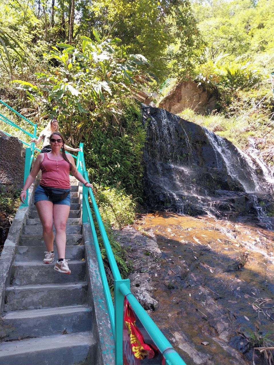 Iemand loopt een betonnen trap af met een groene leuning, omringd door weelderig groen en een kleine waterval.