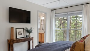 A bedroom with a large bed, a TV mounted on the wall, a wooden console table with a vase and picture, and a window with a view of trees. - Cozy Cottage in Highland Falls Country Club Just 2.5 miles to Downtown Highlands (Highlands)
