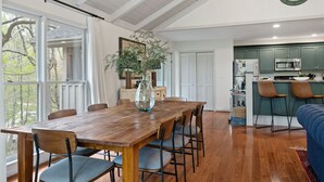 A dining area with a wooden table and chairs, a vase, and a view of trees through large windows. - Cozy Cottage in Highland Falls Country Club Just 2.5 miles to Downtown Highlands (Highlands)