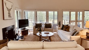 Ein geräumiges Wohnzimmer mit einer großen Couch, einem Couchtisch und einem Fernseher. Große Fenster sorgen für natürliches Licht und einen Ausblick ins Freie.