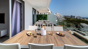 Un balcon moderne avec une table en bois dressée pour les repas, des chaises blanches et une balustrade en verre donnant sur un paysage urbain.