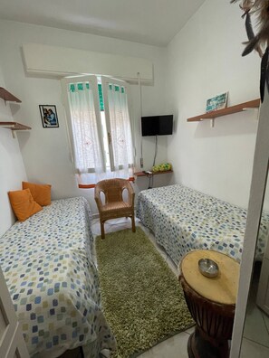 Een kleine, schone kamer met twee enkele bedden, een stoel, een tafeltje, een televisie en een raam met gordijnen.