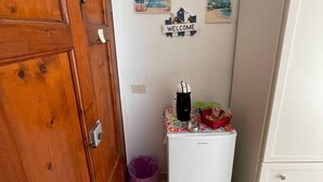 Une petite cuisine équipée d’un réfrigérateur, d’une machine à café et d’une poubelle. Deux panneaux d’accueil sont accrochés au mur, et la porte est en bois.