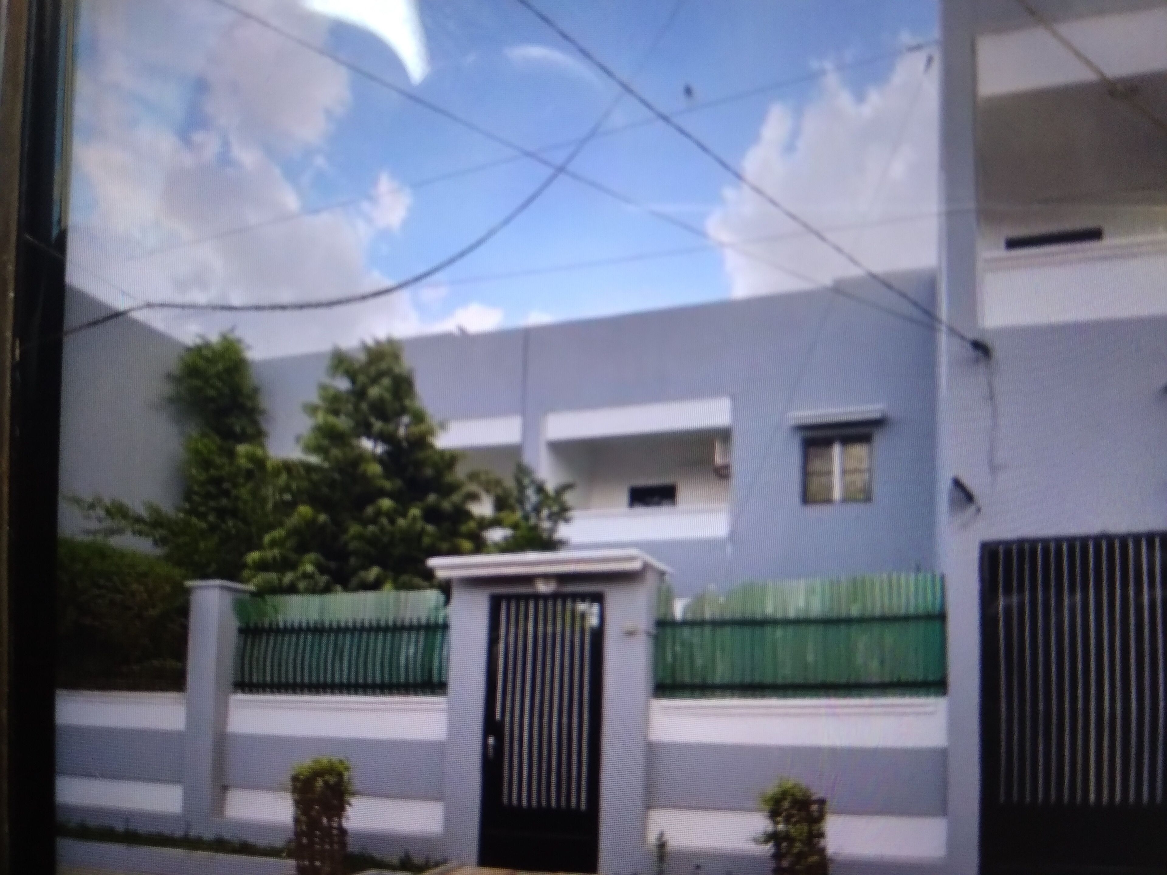 Un edificio con entrada protegida por un portón, una cerca verde y un árbol al frente.