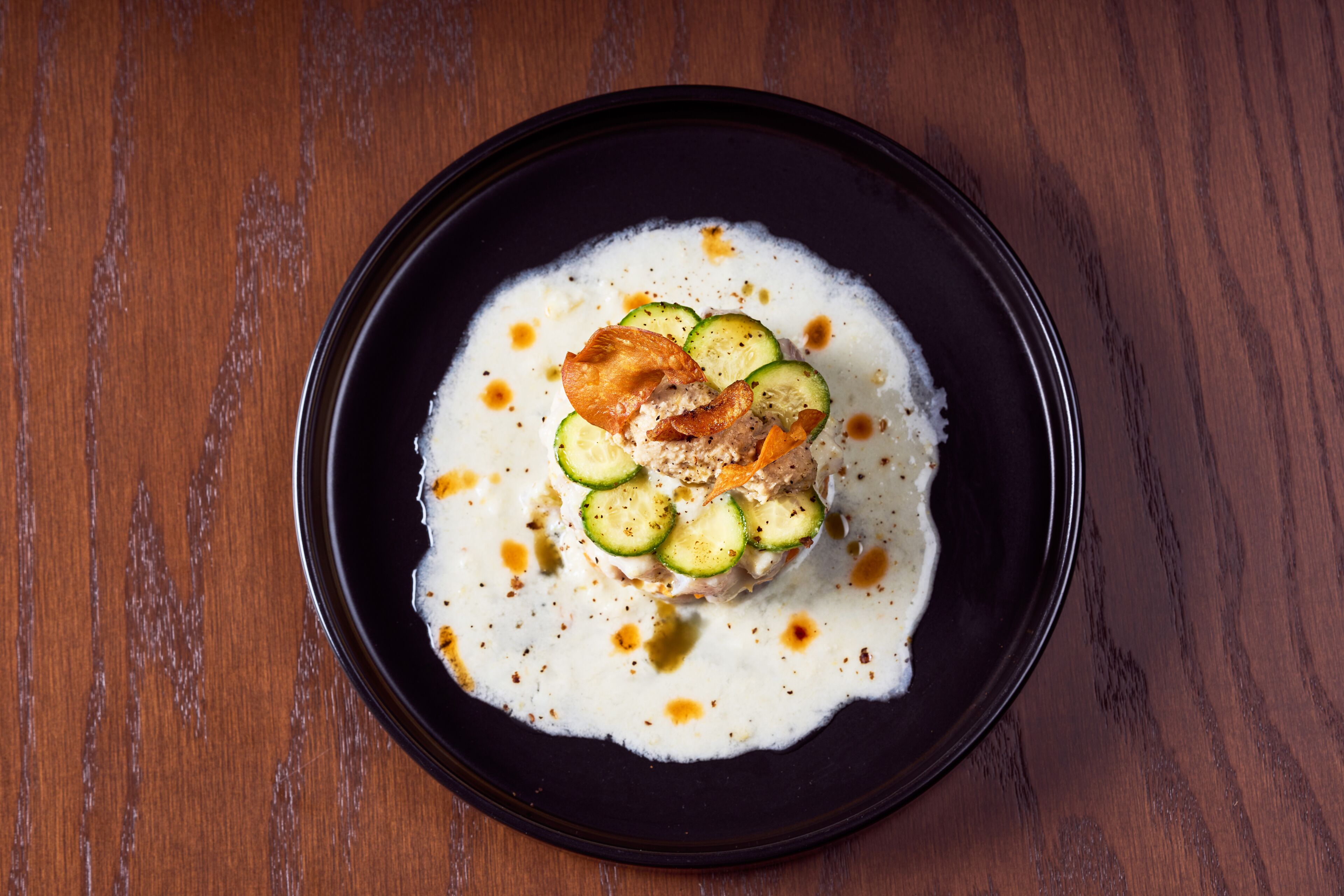 A black plate with a white creamy dish, garnished with cucumber slices and a crispy topping, on a wooden table.