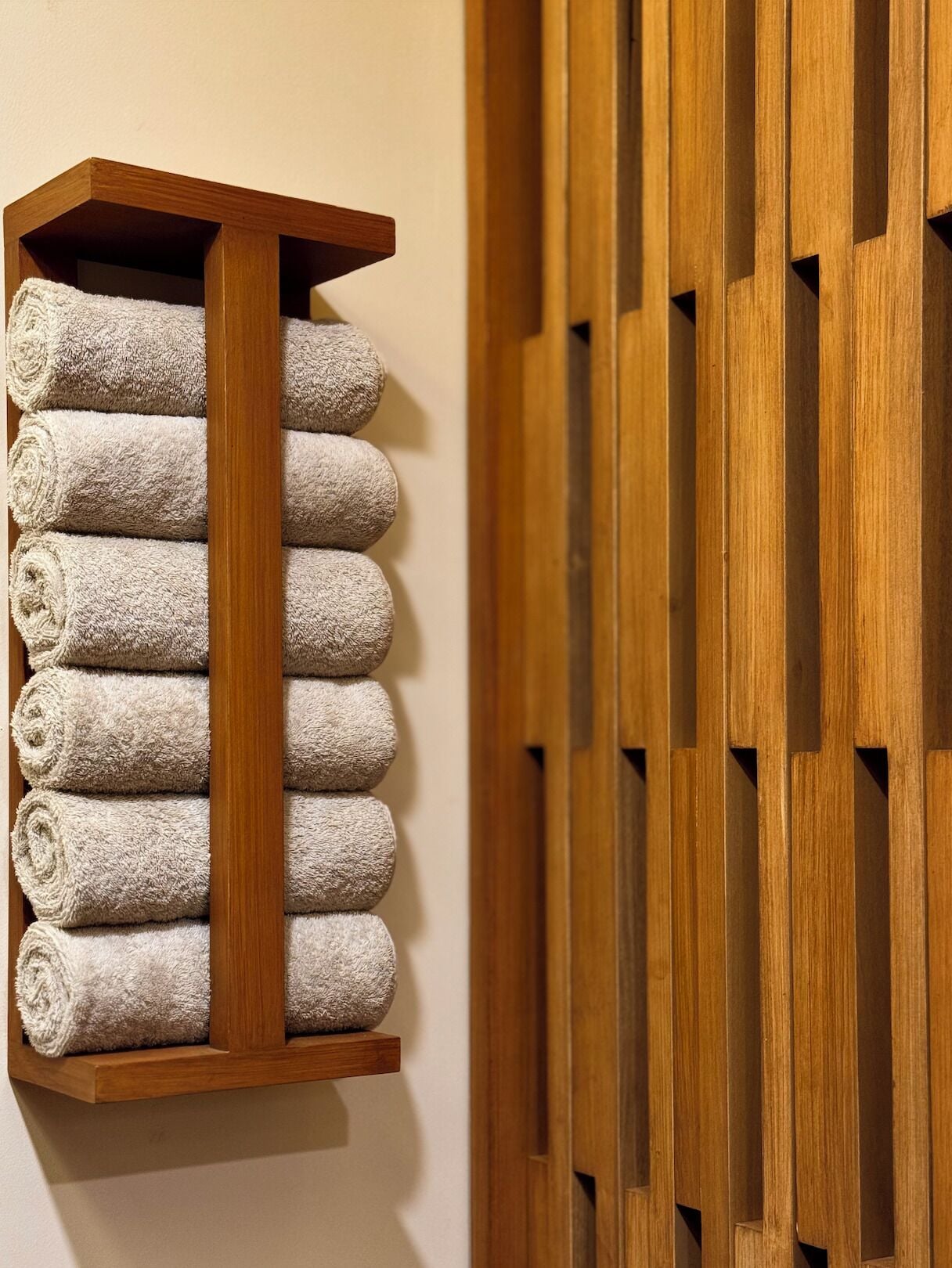 A wooden towel rack with eight neatly rolled towels against a wall.