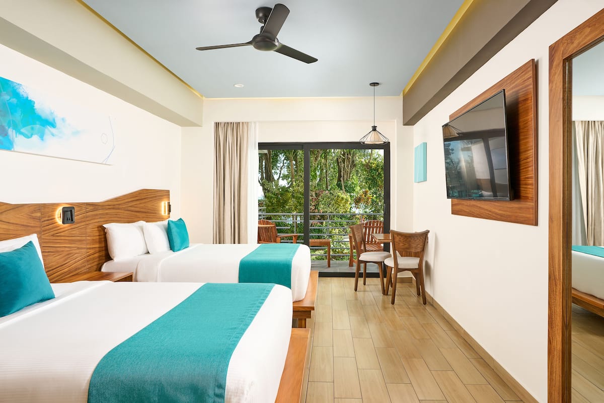 Habitación de hotel con dos camas, ventilador de techo, televisión y balcón con vista a follaje.