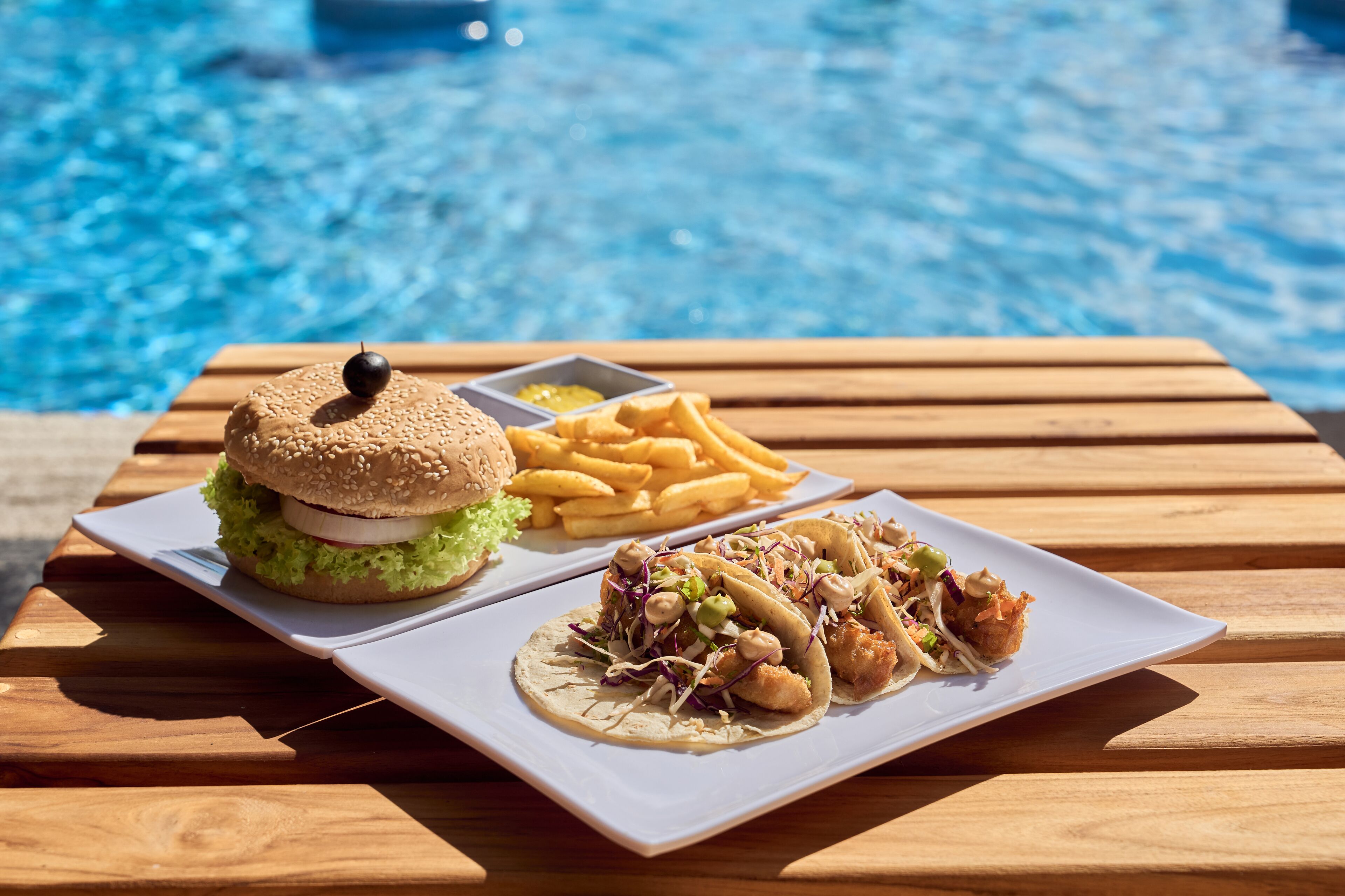 A wooden table with a burger, fries, and tacos by a pool.