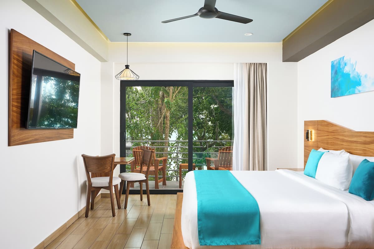 Una habitación de hotel con una cama, televisor montado en la pared, un balcón con sillas y un ventilador de techo.