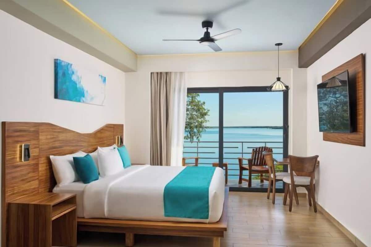 Habitación de hotel con cama, escritorio, silla, ventilador de techo y una ventana grande con vista al mar.