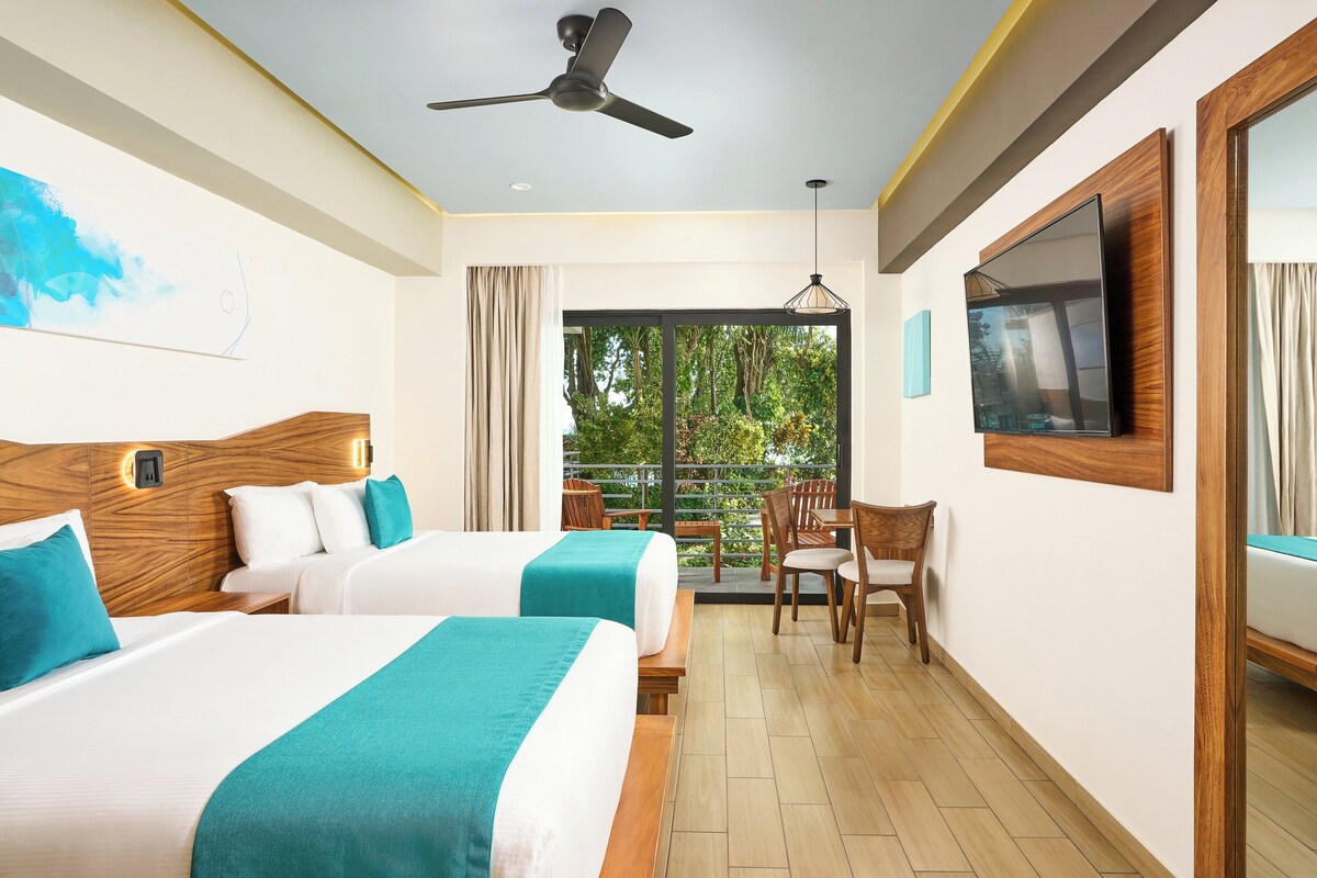 Une chambre d’hôtel avec deux lits, un ventilateur de plafond, une télévision et un balcon donnant sur un paysage verdoyant.