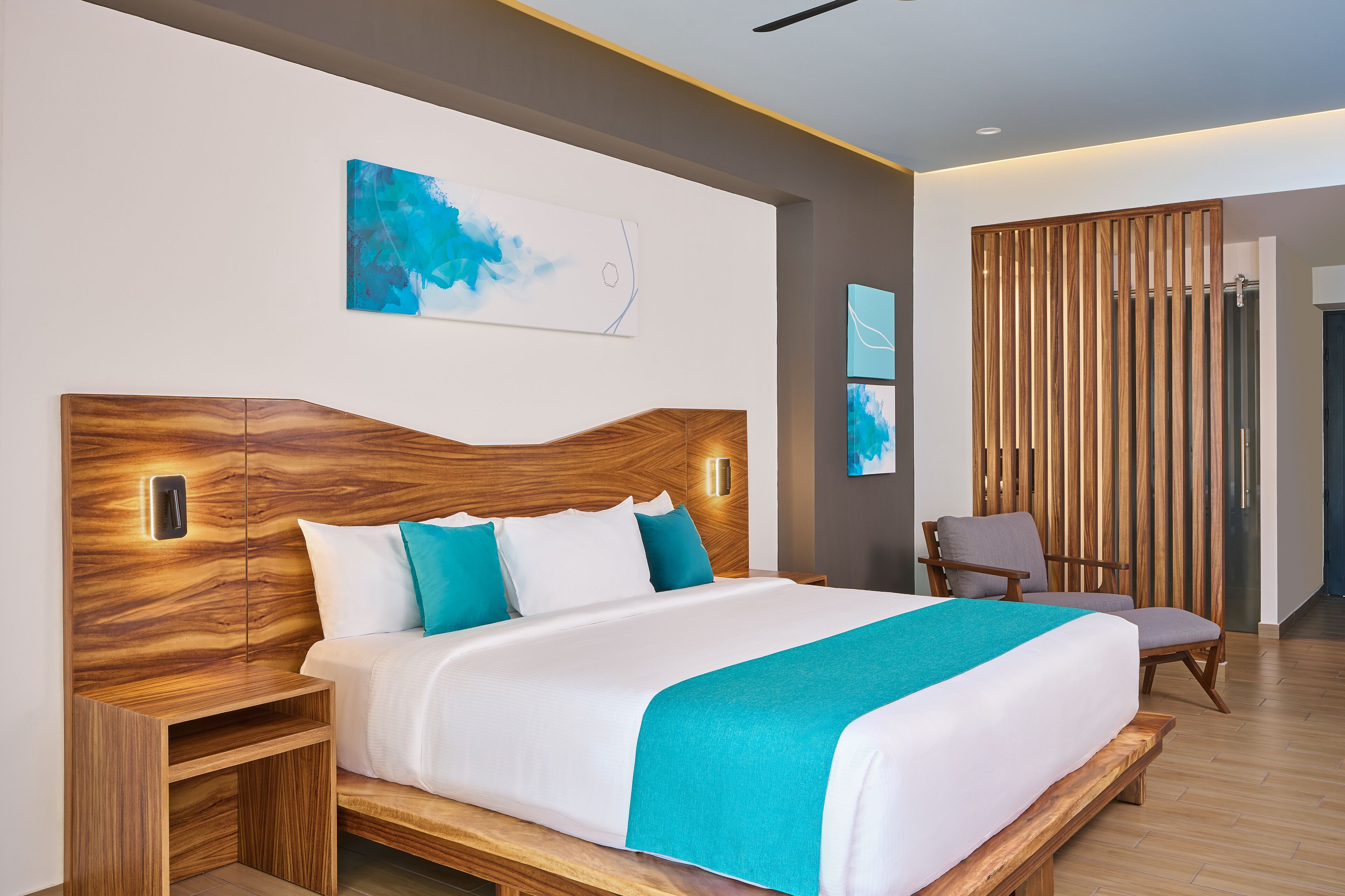 A modern hotel room with a wooden headboard, a large bed with white and teal bedding, a nightstand, and a seating area with a chair and a wooden partition.
