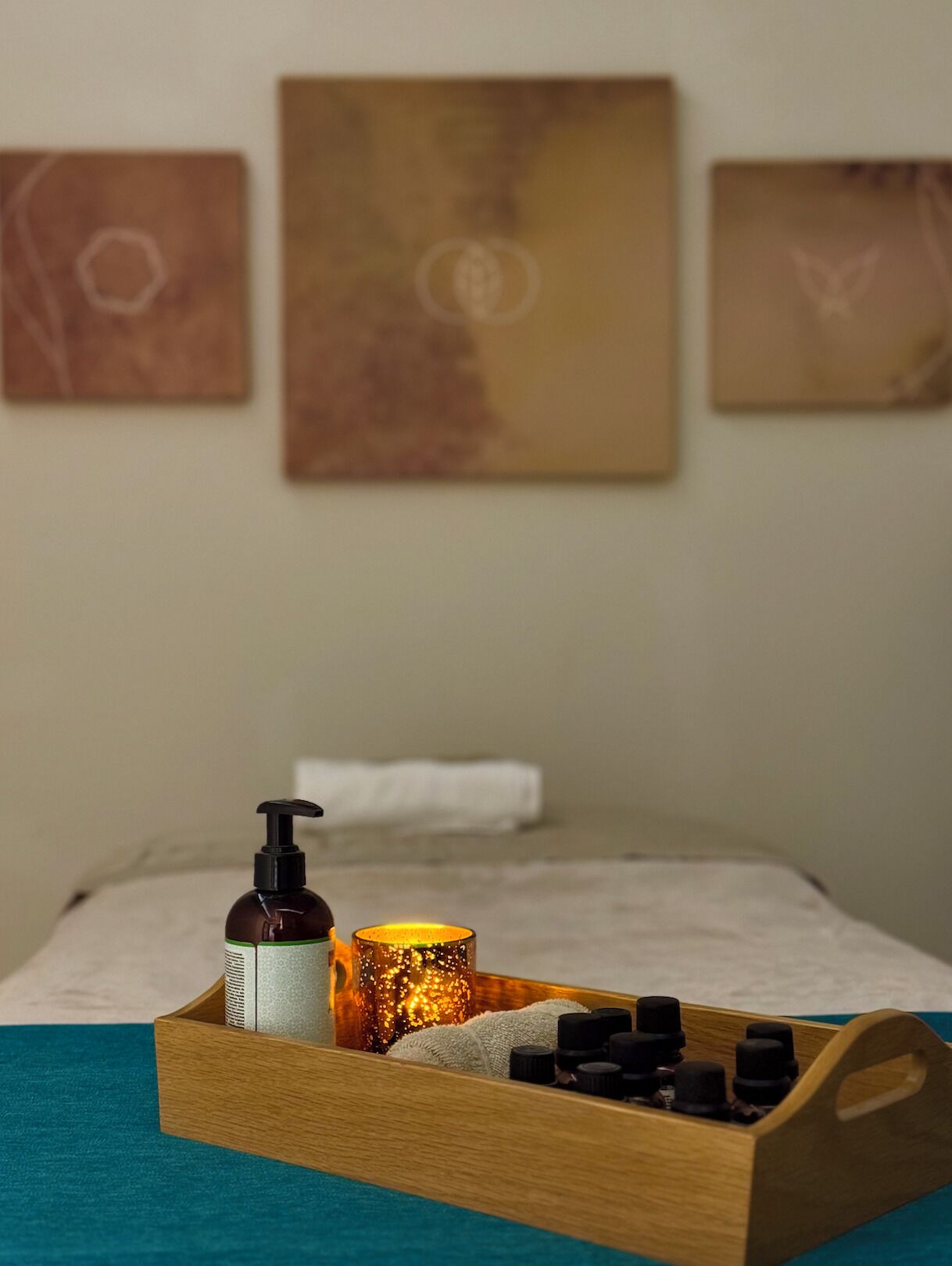A neatly arranged wooden tray on a bed with a bottle of lotion, a candle, and several small bottles, likely containing essential oils, against a backdrop of three abstract paintings on the wall.