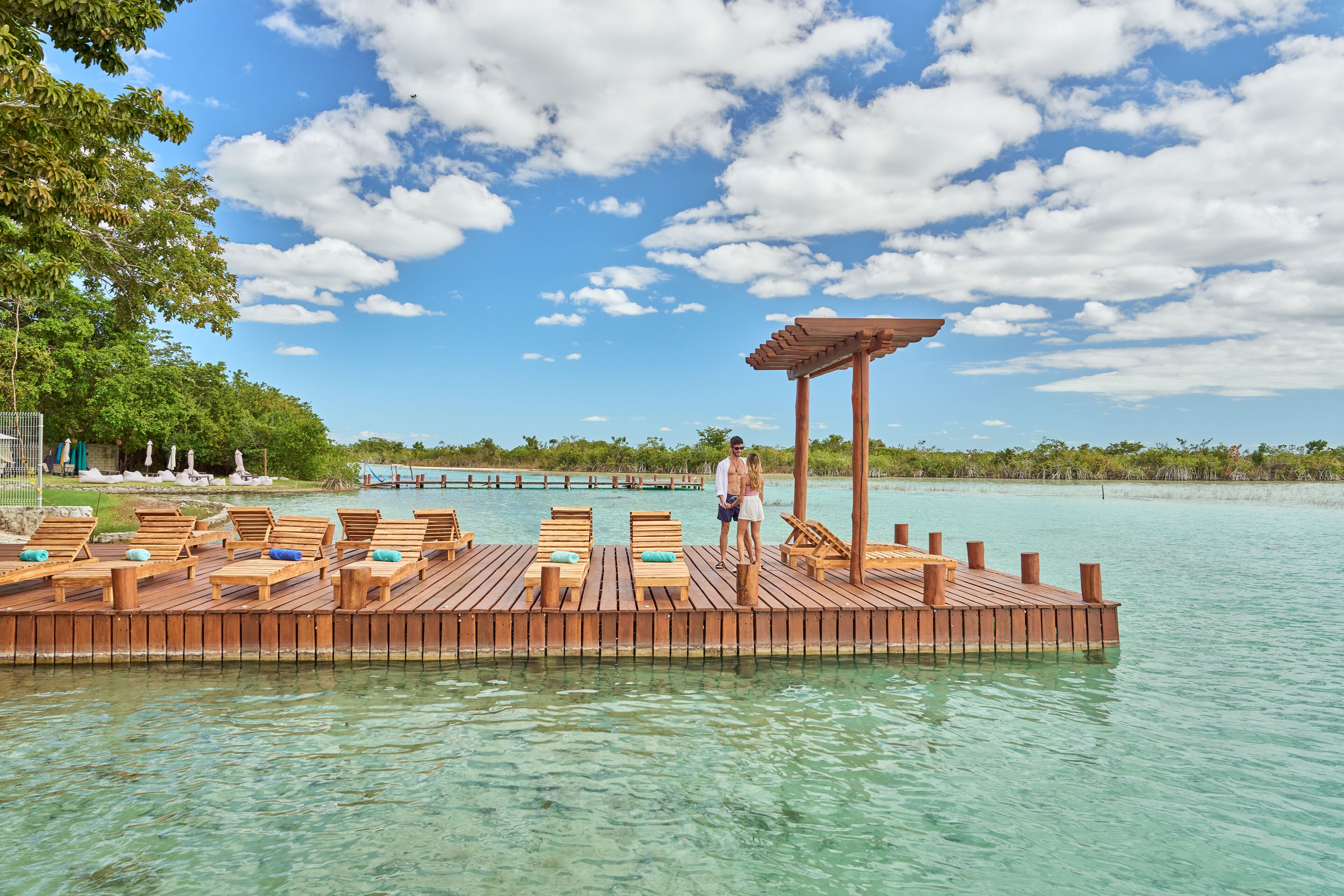 Une plateforme flottante en bois, dotée de chaises longues et d’une pergola, entourée d’une eau cristalline et d’une végétation luxuriante.