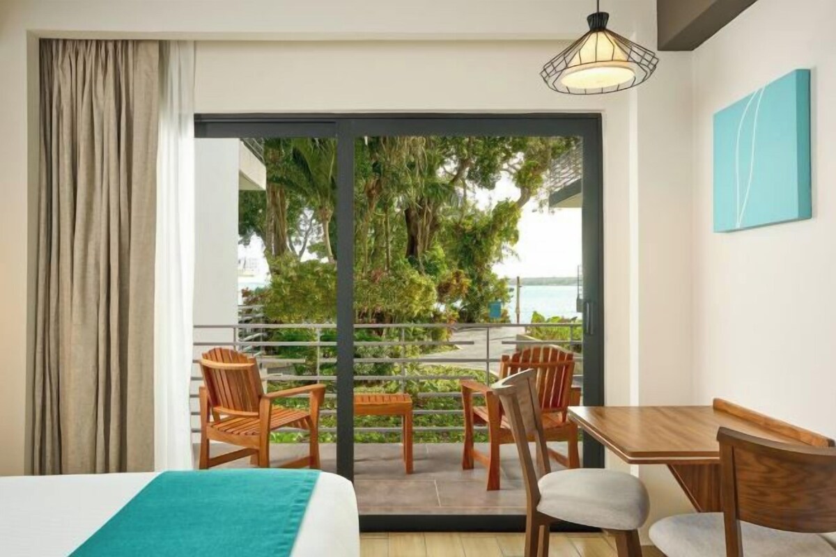 Habitación de hotel con cama, una mesa y sillas, un balcón con vistas a zonas verdes y un cuerpo de agua.