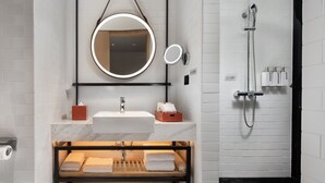 Un baño moderno con un lavabo de mármol, un espejo redondo, un cabezal de ducha de mano y un estante de madera con toallas.