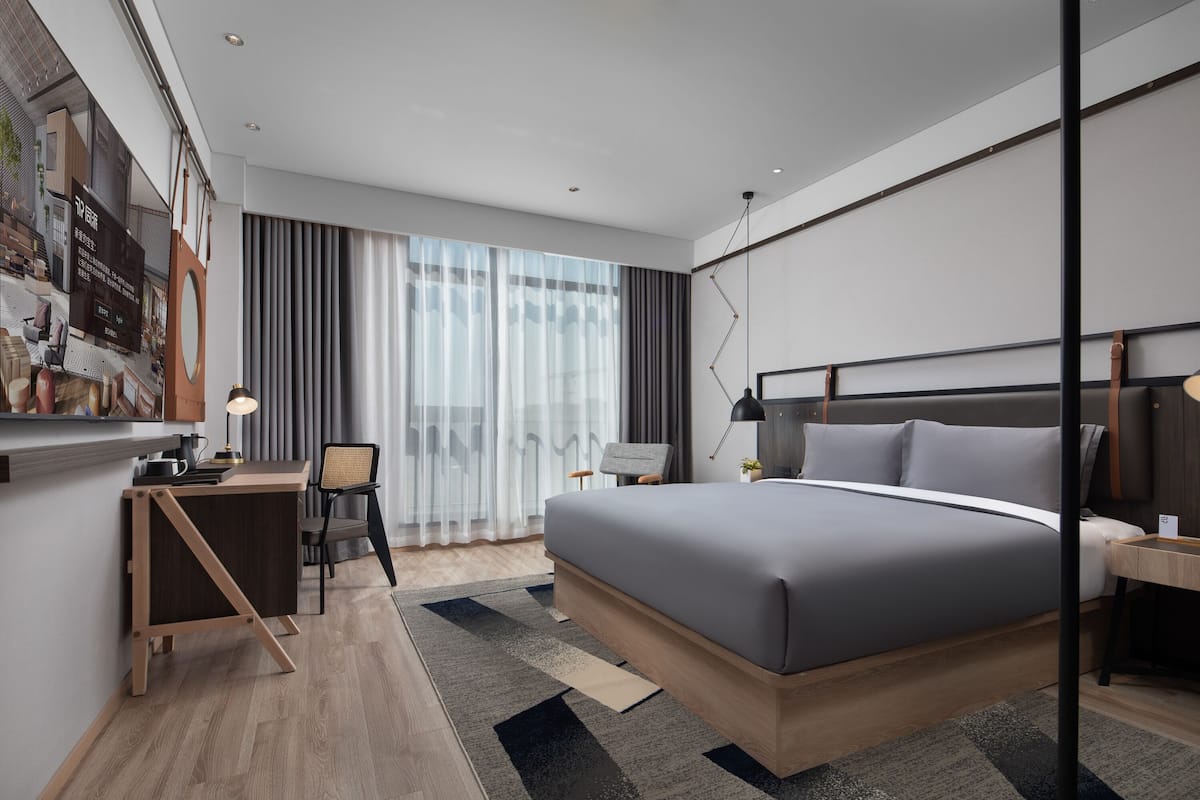 A modern hotel room with a large bed, a desk, and a chair. There is a large window with curtains, a flat-screen TV mounted on the wall, and a small table with a lamp.