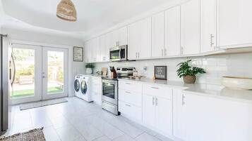 A modern kitchen with white cabinets, a built-in oven, and a microwave. There is a washing machine and a sink in the adjacent area.