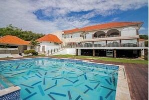 Um hotel com uma piscina grande, um alpendre coberto e um edifício de dois andares com telhado de telhas vermelhas.