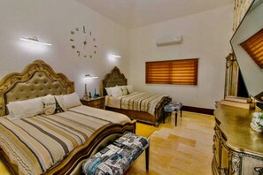 Quarto de hotel com duas camas, um banquinho de madeira, cômoda, relógio e janela com persianas.