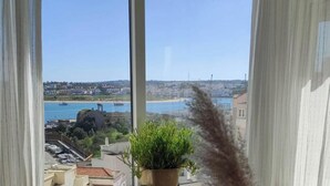 Une vue depuis une fenêtre, avec une plante en pot au premier plan, donnant sur une zone côtière avec des bâtiments et une étendue d’eau.