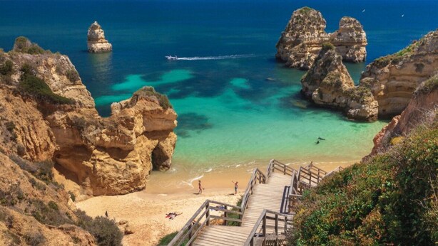 Une plage côtière bordée de formations rocheuses, aux eaux turquoise et limpides, et dotée d’une promenade en bois menant à la rive.