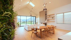 A modern living room with a wooden dining table and chairs, a white sofa, and large windows offering a view of the outdoors.