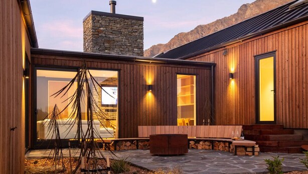 A modern house with a stone chimney, wooden exterior, and a stone patio with outdoor seating.