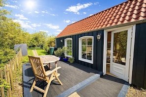 Une terrasse en bois avec une table et des chaises, un patio couvert et un bâtiment doté d’un toit recouvert de tuiles rouges et d’une porte.
