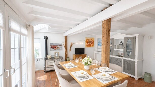 Un comedor con una mesa de madera dispuesta para cuatro personas, rodeada de sillas. Se observa una zona de cocina con armarios y un televisor al fondo.
