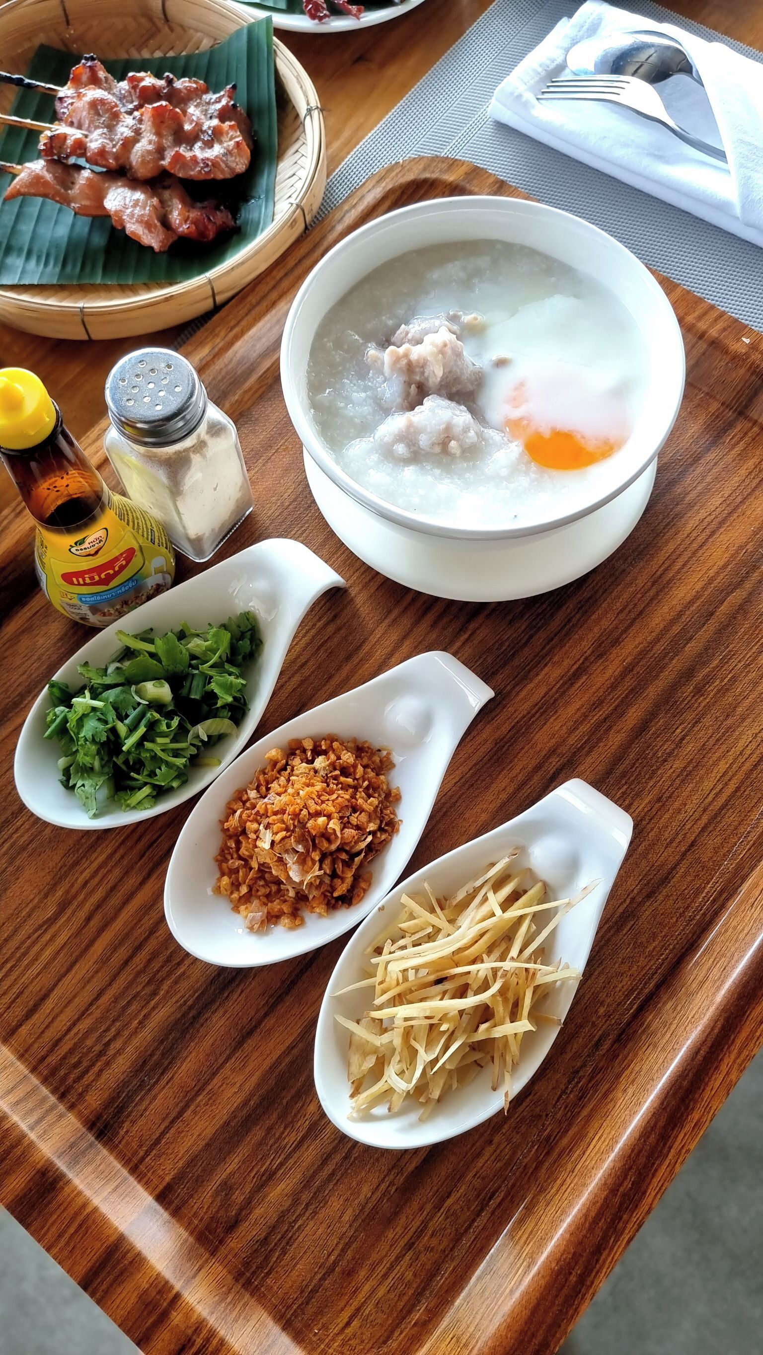 A wooden table with a bowl of soup, skewers of meat, chopped green onions, fried shallots, and a bottle of soy sauce.