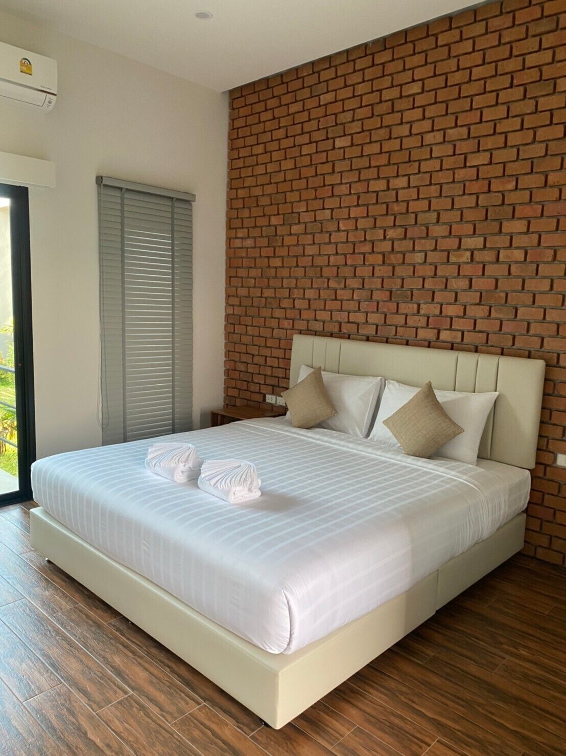 A neatly made bed with white linens and two towels, a brick-patterned wall, and a wooden floor.