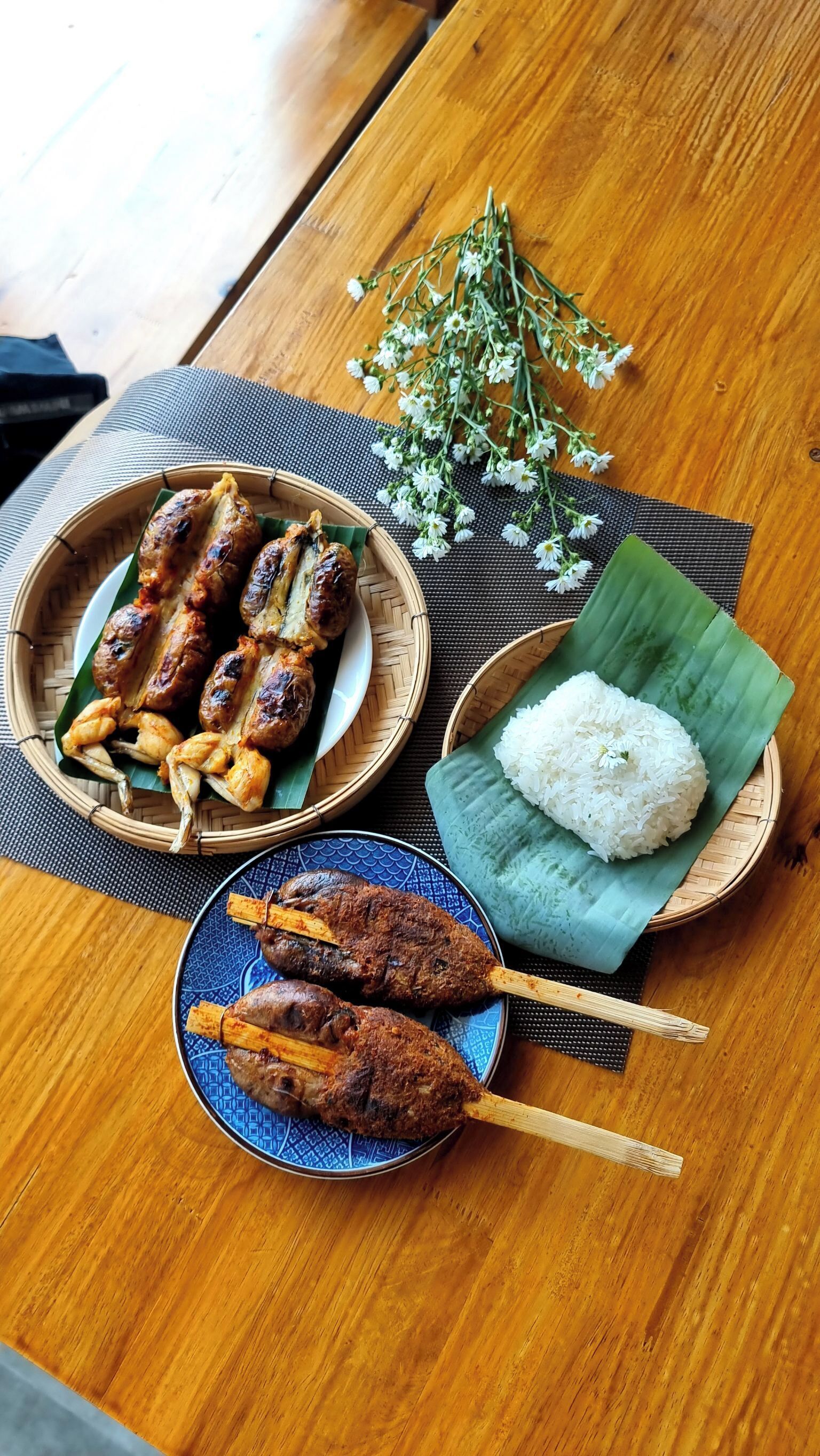 A wooden table with a plate of grilled meat skewers, a bowl of white rice on a banana leaf, and a decorative branch of white flowers.