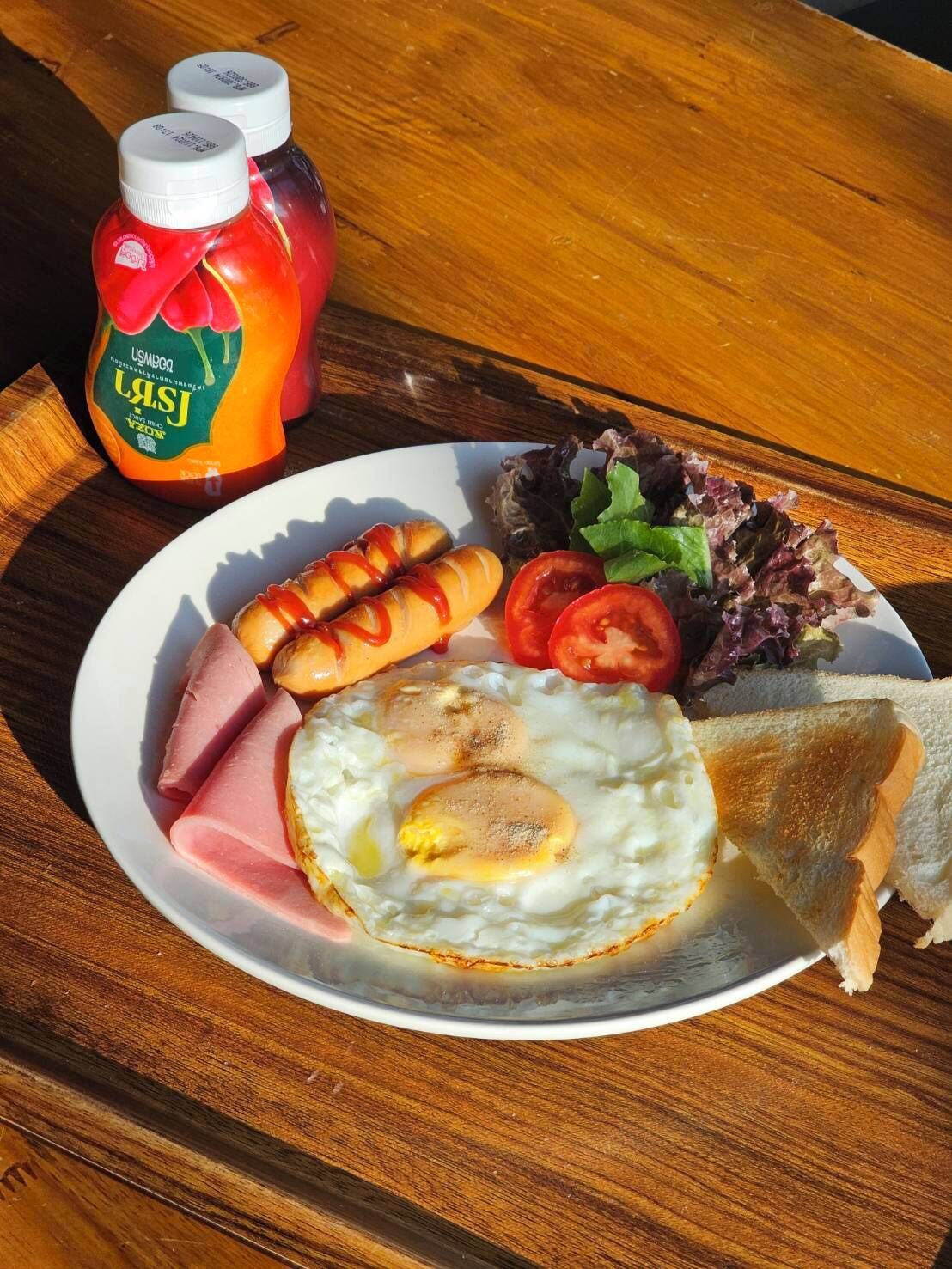 A plate with eggs, sausages, ham, tomatoes, lettuce, and toast on a wooden table.