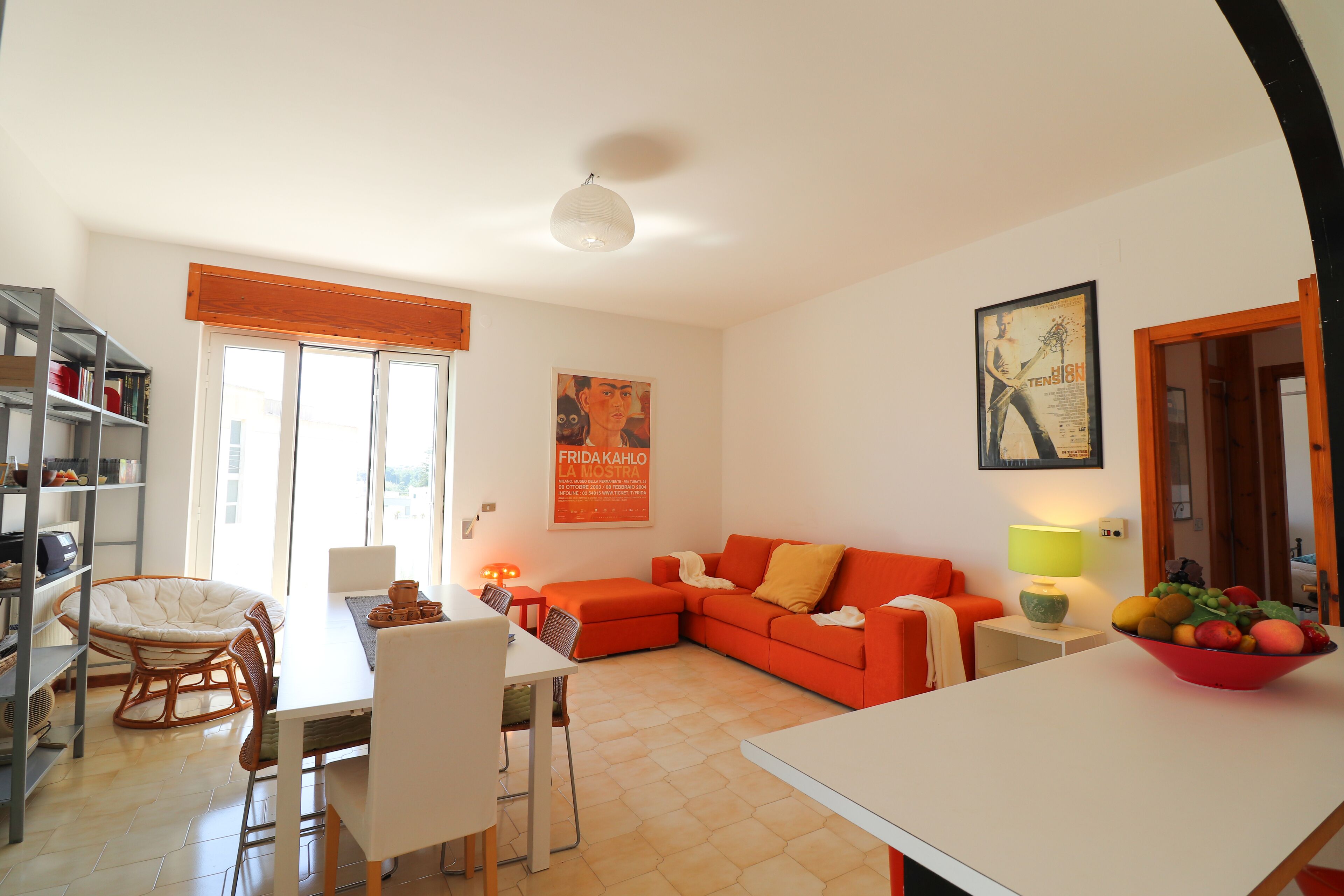 Un salon moderne comprenant un coin repas, un canapé et une table avec des chaises. On y trouve également une bibliothèque, une fenêtre avec des stores et un tableau accroché au mur.