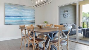 Ein Essbereich mit Holztisch und Stühlen, einem Gemälde mit Strandmotiv und Blick auf eine Außenbestuhlung durch eine Glastür.