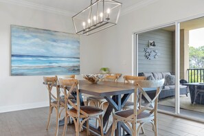 Ein Essbereich mit Holztisch und Stühlen, einem Gemälde mit Strandmotiv und Blick auf eine Außenbestuhlung durch eine Glastür.