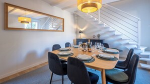 Uma sala de jantar com uma mesa grande posta para seis pessoas, um espelho na parede e uma escada ao fundo.