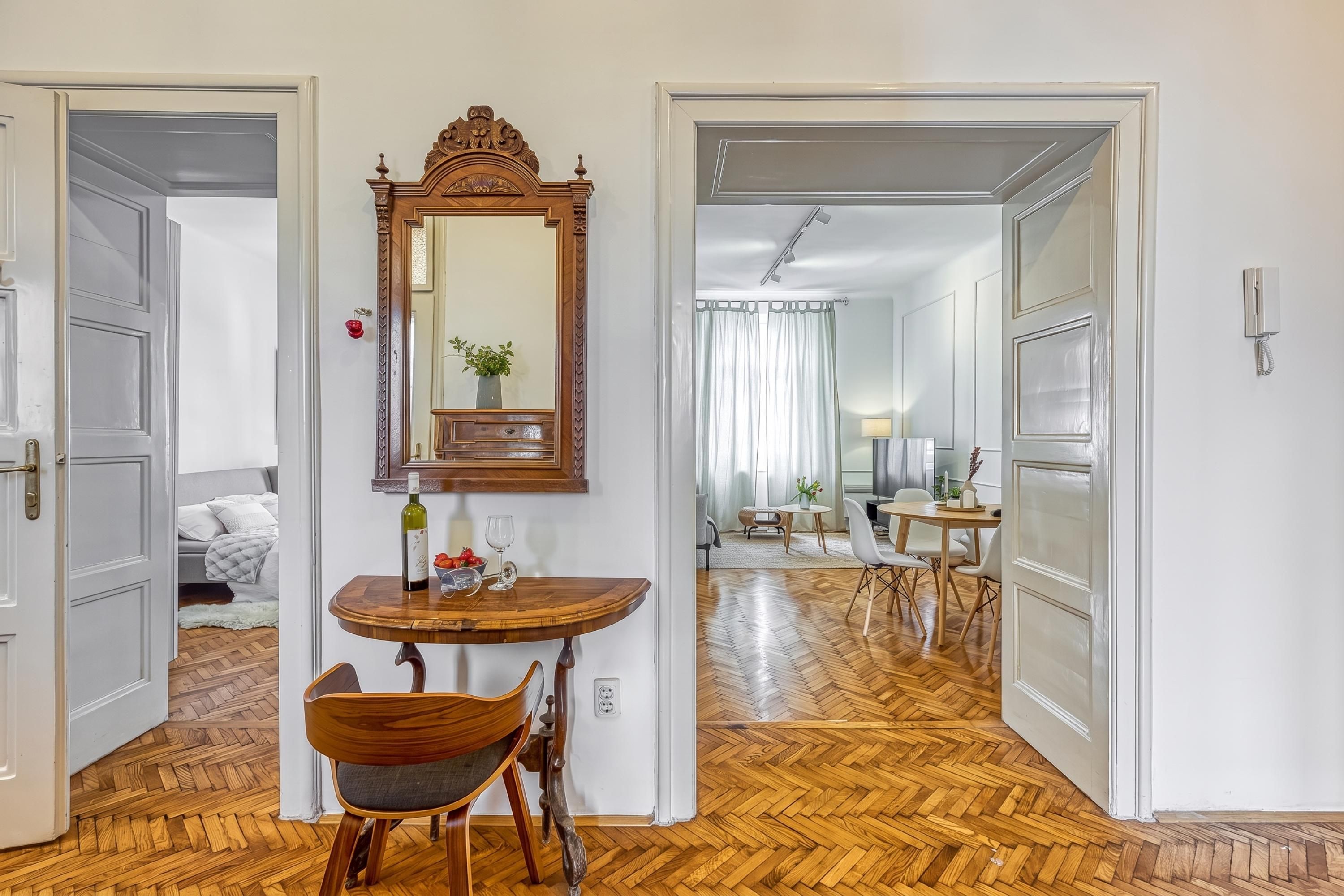 A room with a wooden floor, a dining table with chairs, a mirror, and a bed with pillows.
