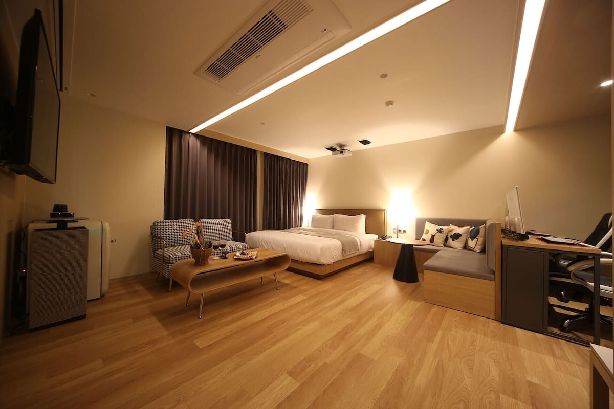 A modern hotel room with a bed, a sofa, a desk with a computer, and a TV.