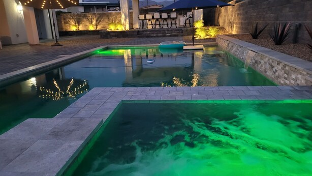 En pool med ett litet vattenfall, omgiven av en uteplats med utemöbler och ljusslingor.
