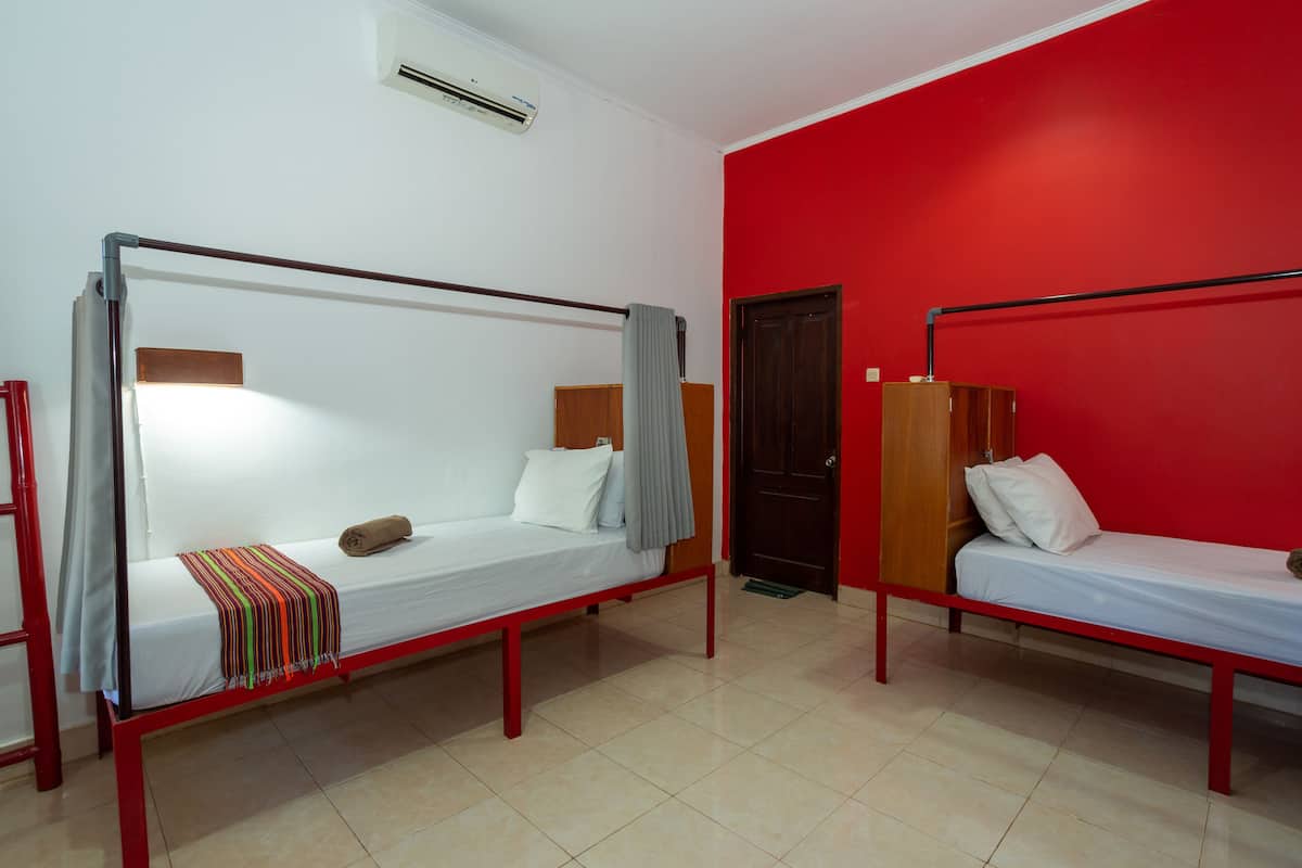 Una habitación con dos camas individuales, cada una con cabecero de madera y ropa de cama blanca. Una pared es roja y la otra blanca, con un equipo de aire acondicionado.