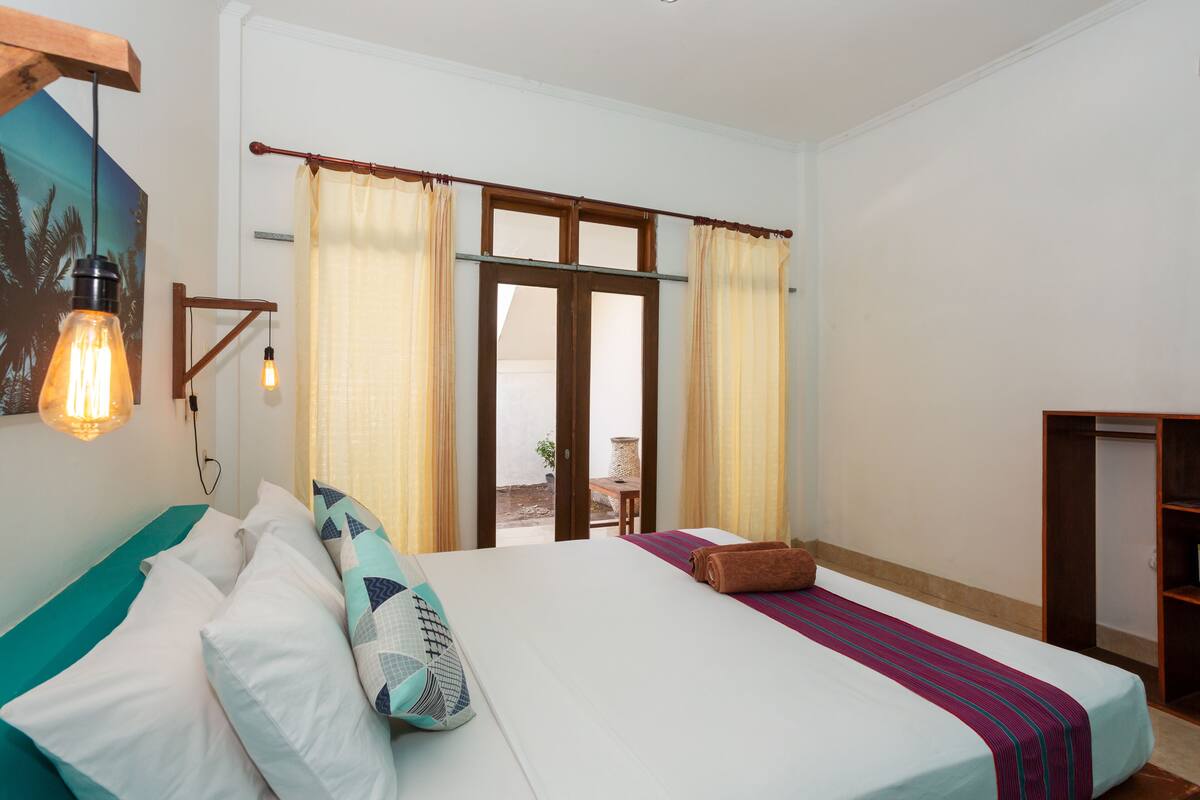 Un dormitorio con cama, almohadas, cabecero de madera, ventana con cortinas y vistas al exterior.