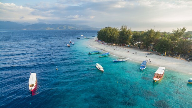 Una playa con aguas turquesas cristalinas, varias embarcaciones ancladas cerca de la costa y una playa de arena con árboles y sombrillas.