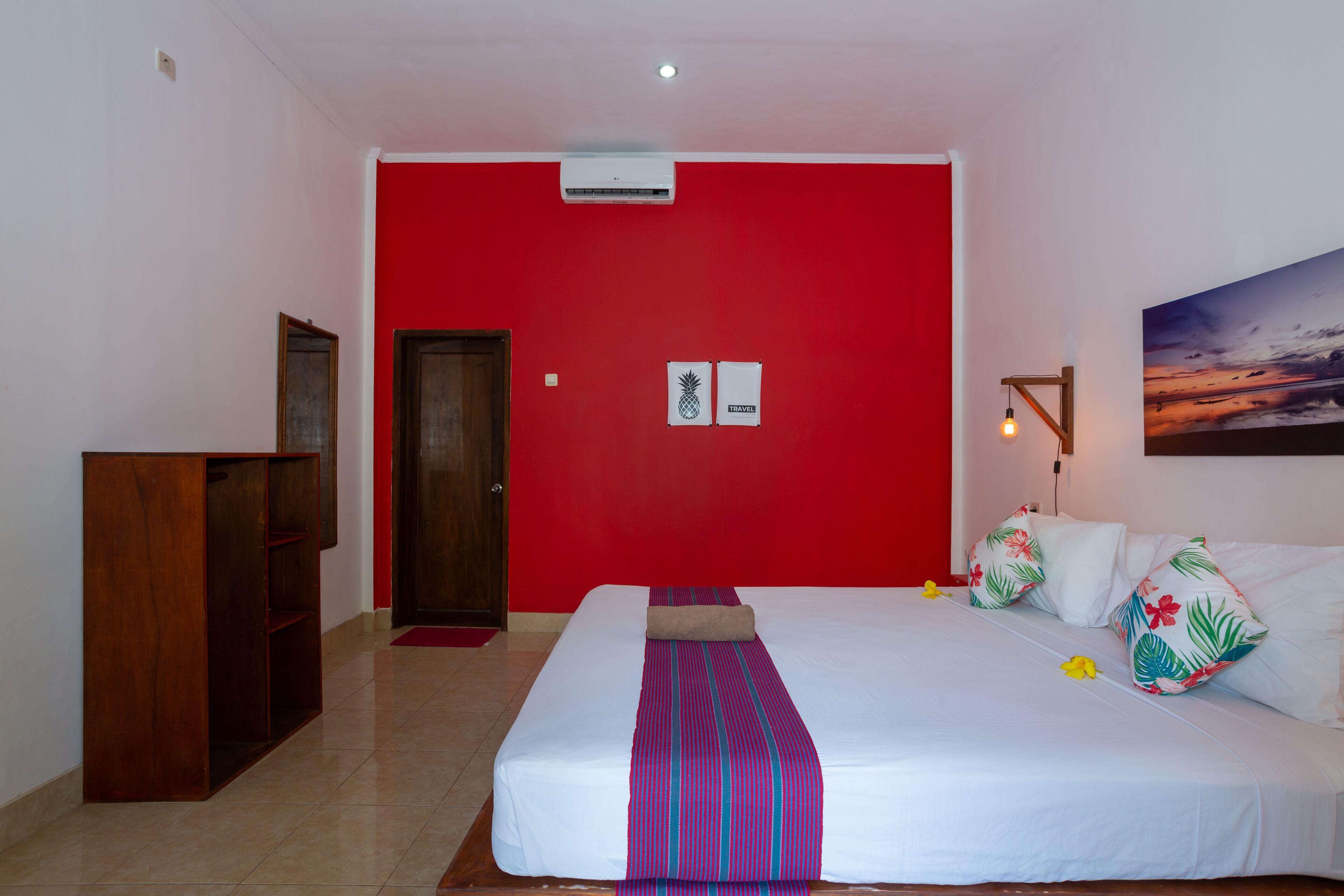A hotel room with a red accent wall, a bed with a striped bedspread, tropical-themed pillows, and a painting of a sunset.