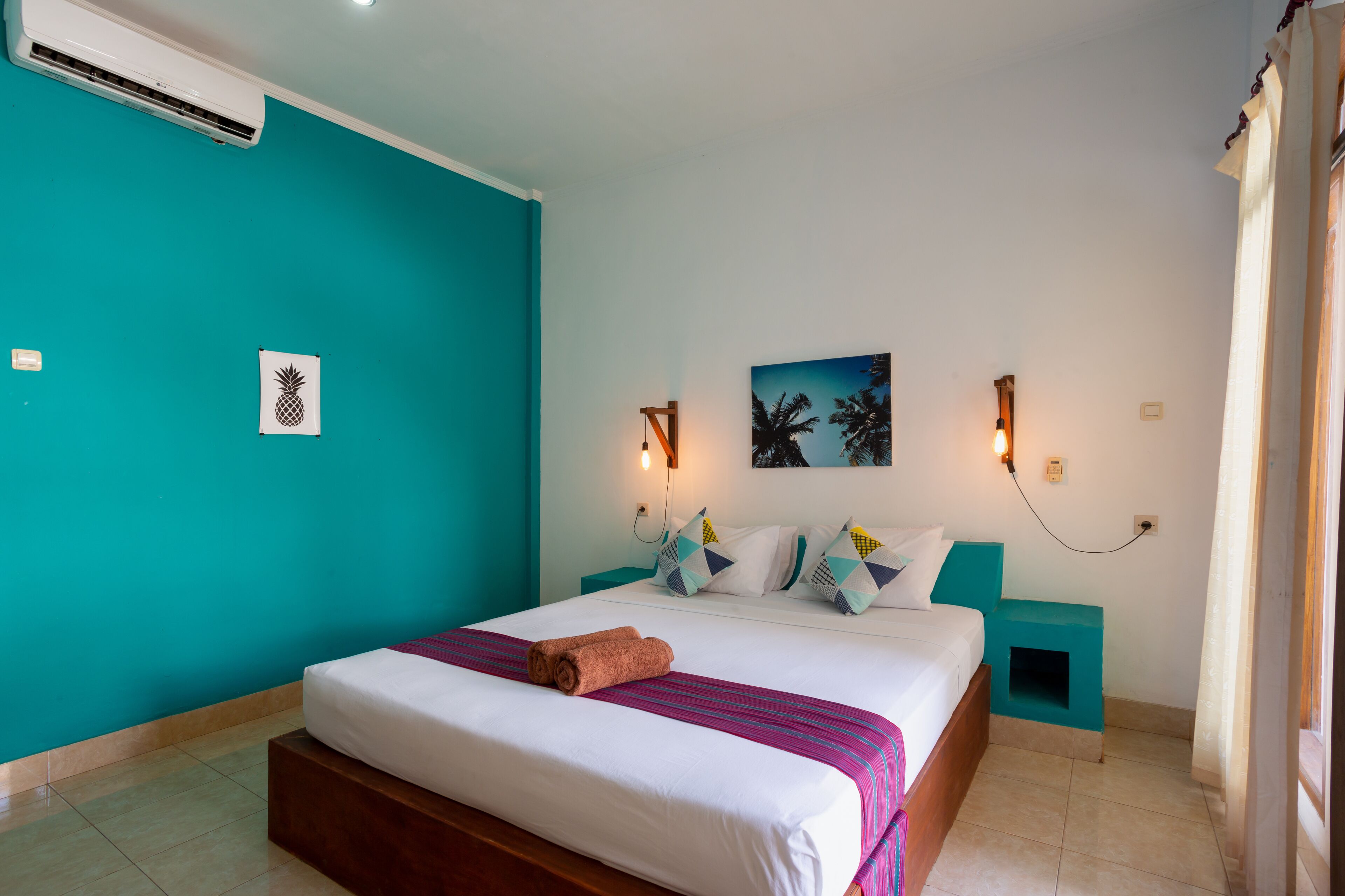 A hotel room with a bed, teal walls, a painting, and a wall-mounted air conditioner.