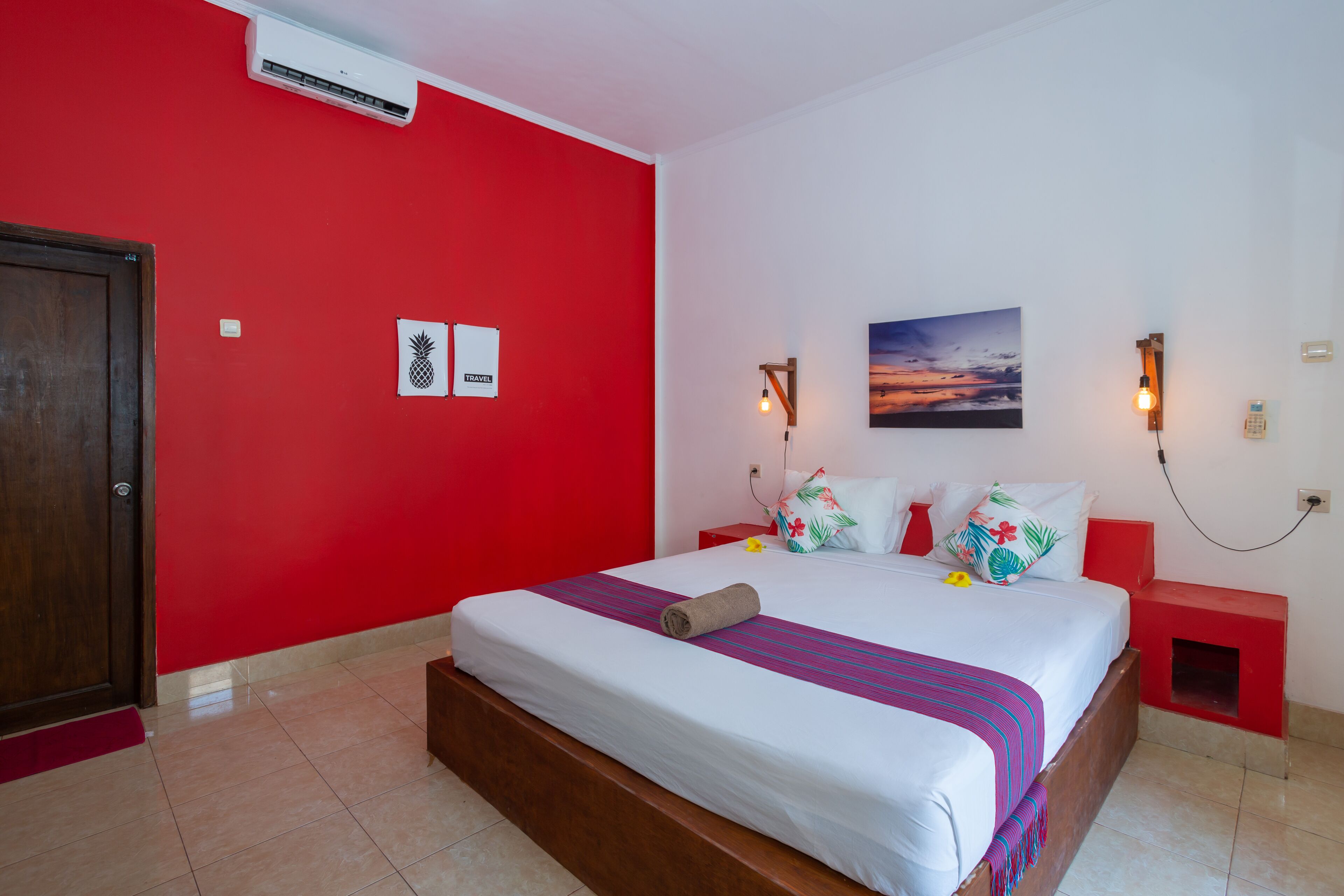 A hotel room with a red wall, a bed with a white and purple striped blanket, a wooden headboard, a nightstand with a red lamp, and a painting of a sunset.