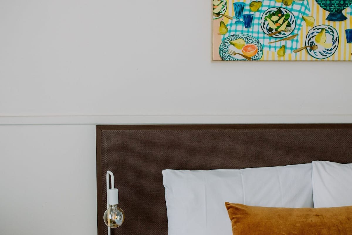 A neatly made bed with white and brown bedding, a small round side table with a lamp, and a painting of fruit and flowers on the wall.