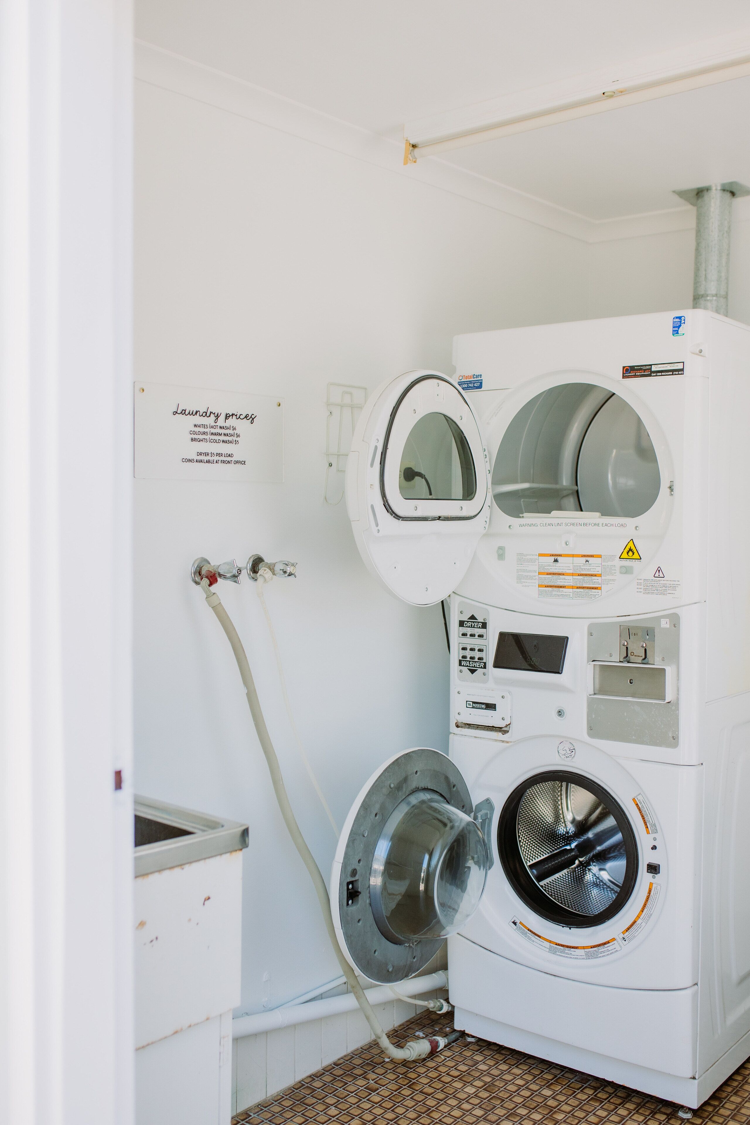 Une buanderie équipée de deux machines à laver et d’un sèche-linge, un panneau indiquant les tarifs de lavage et une étiquette d’avertissement de sécurité.